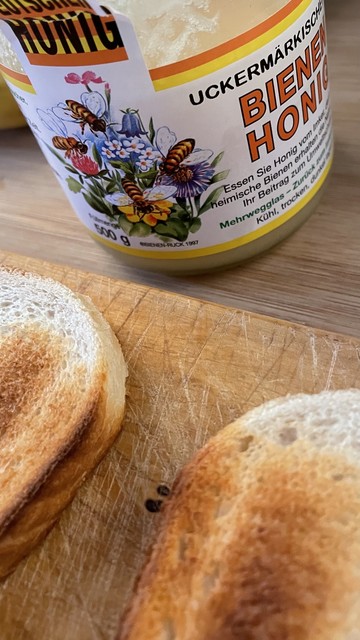 Ein Glas Honig mit Blumen- und Bienenmotiven auf dem Etikett, das hinter zwei getoasteten Brotscheiben auf einem hölzernen Schneidebrett steht.