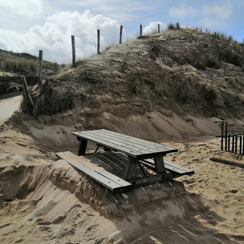 Eine Pikniktafel (Holztisch-/Bankkombination) in den Dünen, unter die so viel Sand geschoben oder geschaufelt wurde, dass man nicht mehr dran sitzen kann, weil die Beine keinen Platz haben. 