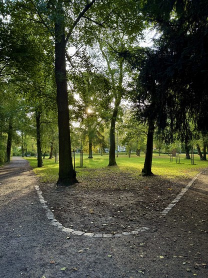 Das Bild zeigt einen schönen Park an einem sonnigen Tag. Im Vordergrund sieht man einen gepflasterten Weg, der sich in zwei Richtungen teilt. Der Weg ist von hohen, alten Bäumen gesäumt, deren Blätterdach ein grünes Dach über dem Weg bildet.

Durch die Bäume hindurch fällt warmes Sonnenlicht, das einzelne Stellen des Rasens und des Weges erhellt. Dies erzeugt ein reizvolles Spiel von Licht und Schatten.

Im Hintergrund erstreckt sich eine offene Rasenfläche, auf der vereinzelt jüngere Bäume stehen. Die gesamte Szene vermittelt einen friedlichen und einladenden Eindruck eines gepflegten Stadtparks.

Die Atmosphäre wirkt ruhig und entspannend, ideal für einen Spaziergang oder eine Pause im Grünen. Das saftige Grün der Bäume und des Rasens kontrastiert angenehm mit dem grauen Weg, was dem Bild eine harmonische Balance verleiht.​​​​​​​​​​​​​​​​