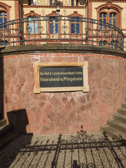 durch ein schmiedeeisernes Tor, welches noch einen Schatten wirft, eine schwarze Tafel an einer rötlichen  Natursteinmauer fotografiert.
auf der Tafel steht:
Der Rat der Landeshauptstadt Halle
Feierabend- u. Pflegeheim.

Halle ist schon lange nicht mehr Landeshauptstadt und betreibt IMHO auch keine eigenen Pflegeheime...